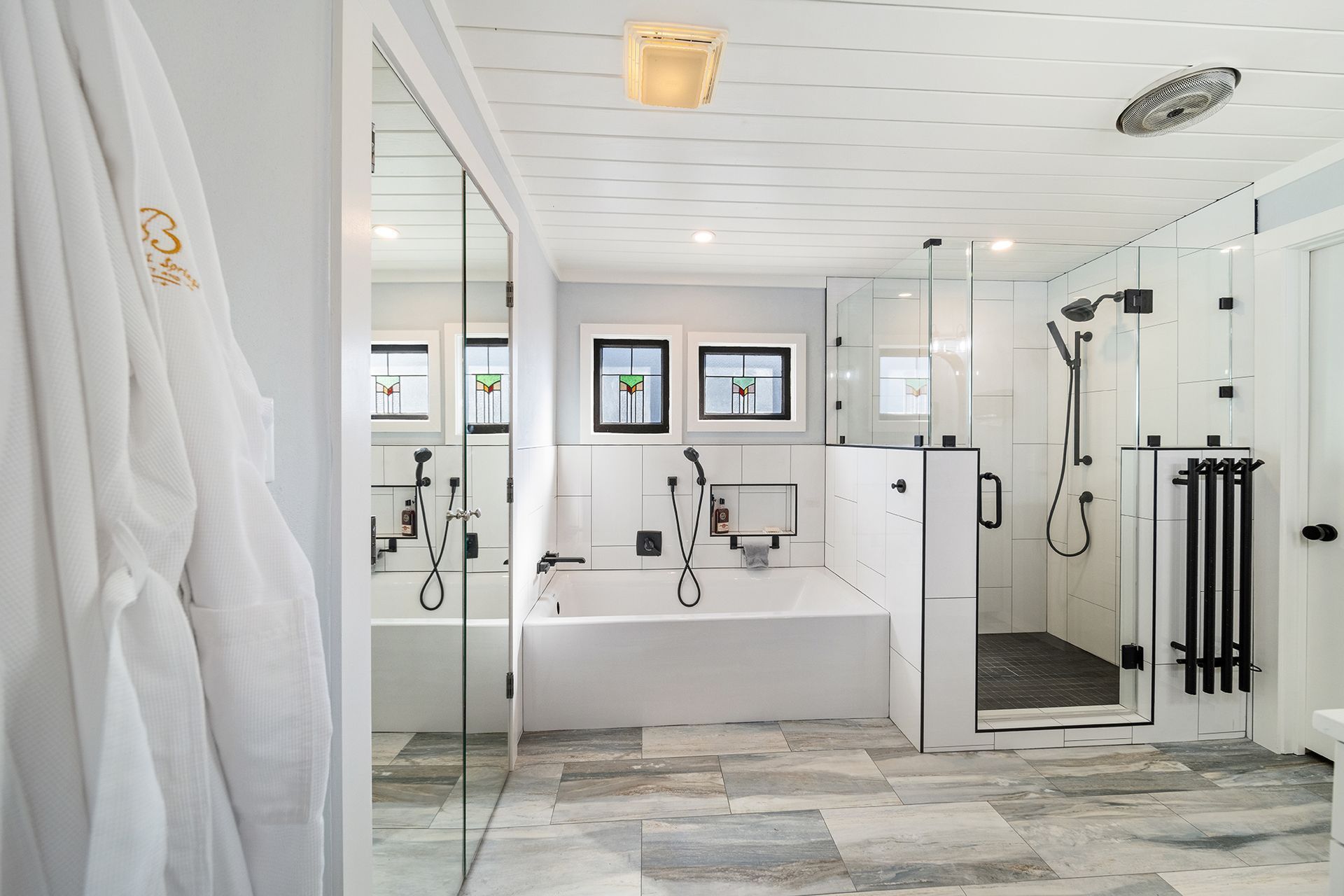 White and modern bathroom with soaking tub, walk-in shower, and a robe hanging on the door. Gray tiled floor.