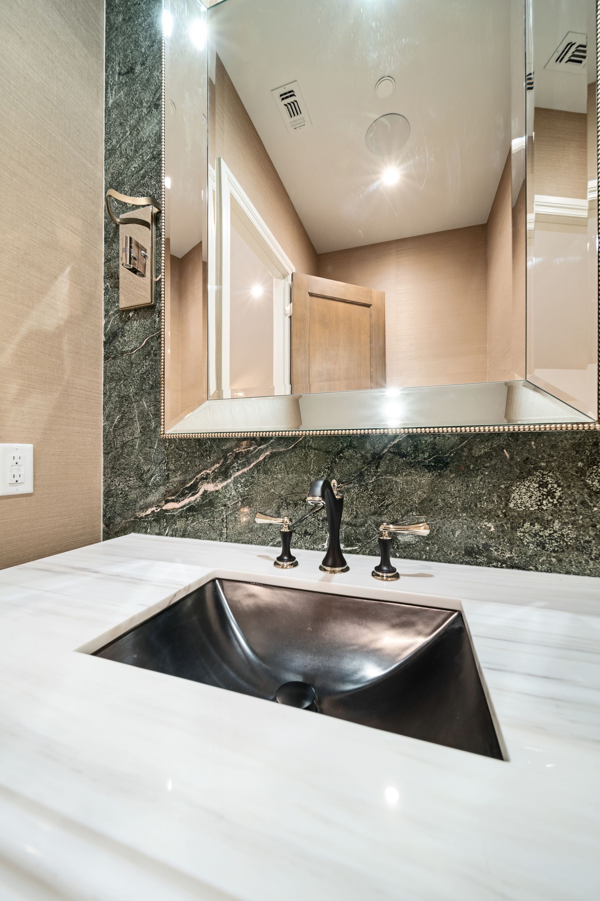 Bathroom sink with black basin and dark fixtures against a marble countertop. Mirror reflects a door and light fixtures.