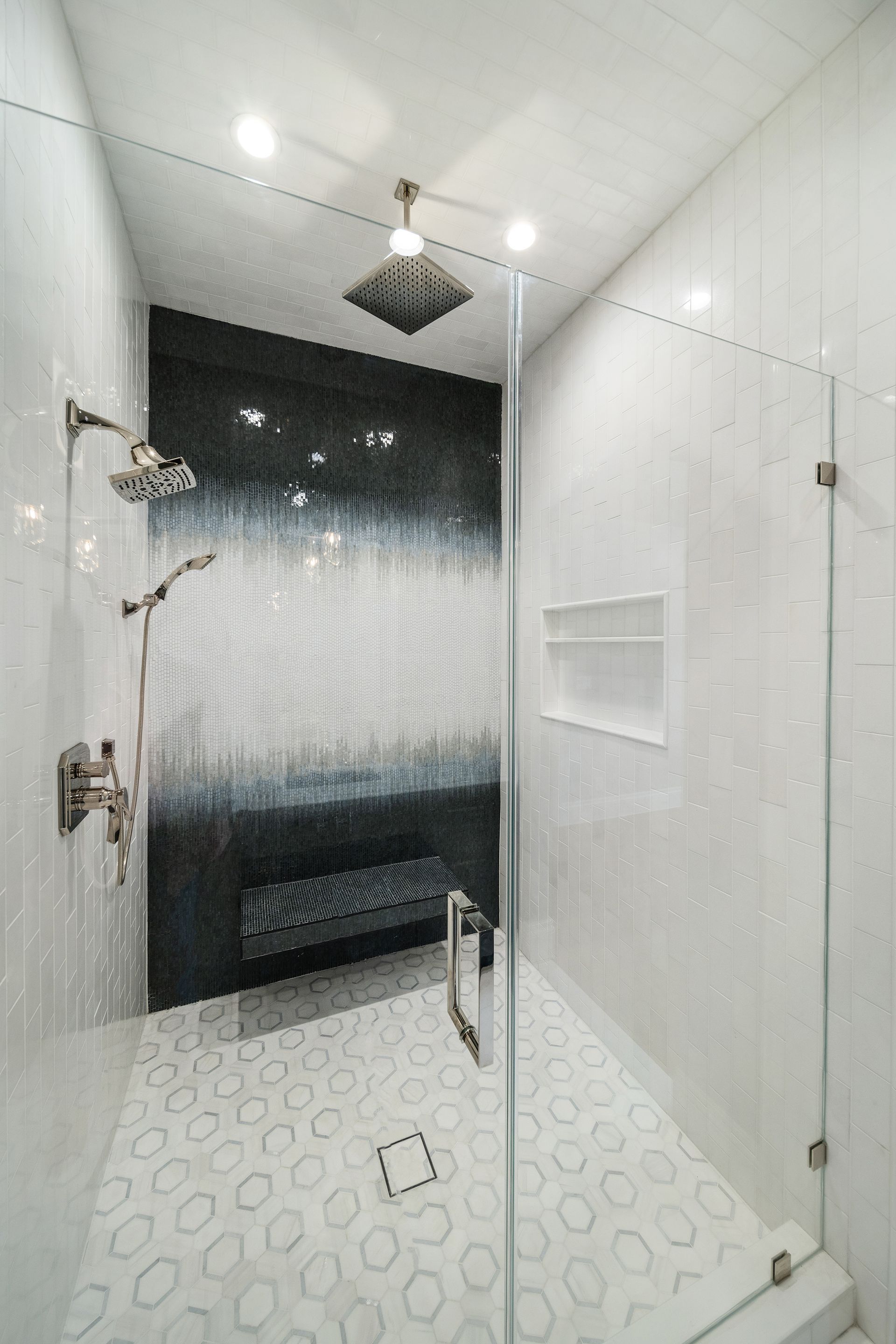 Modern shower with white and gray tiles. Dark blue accent wall, glass door, and recessed shelf.