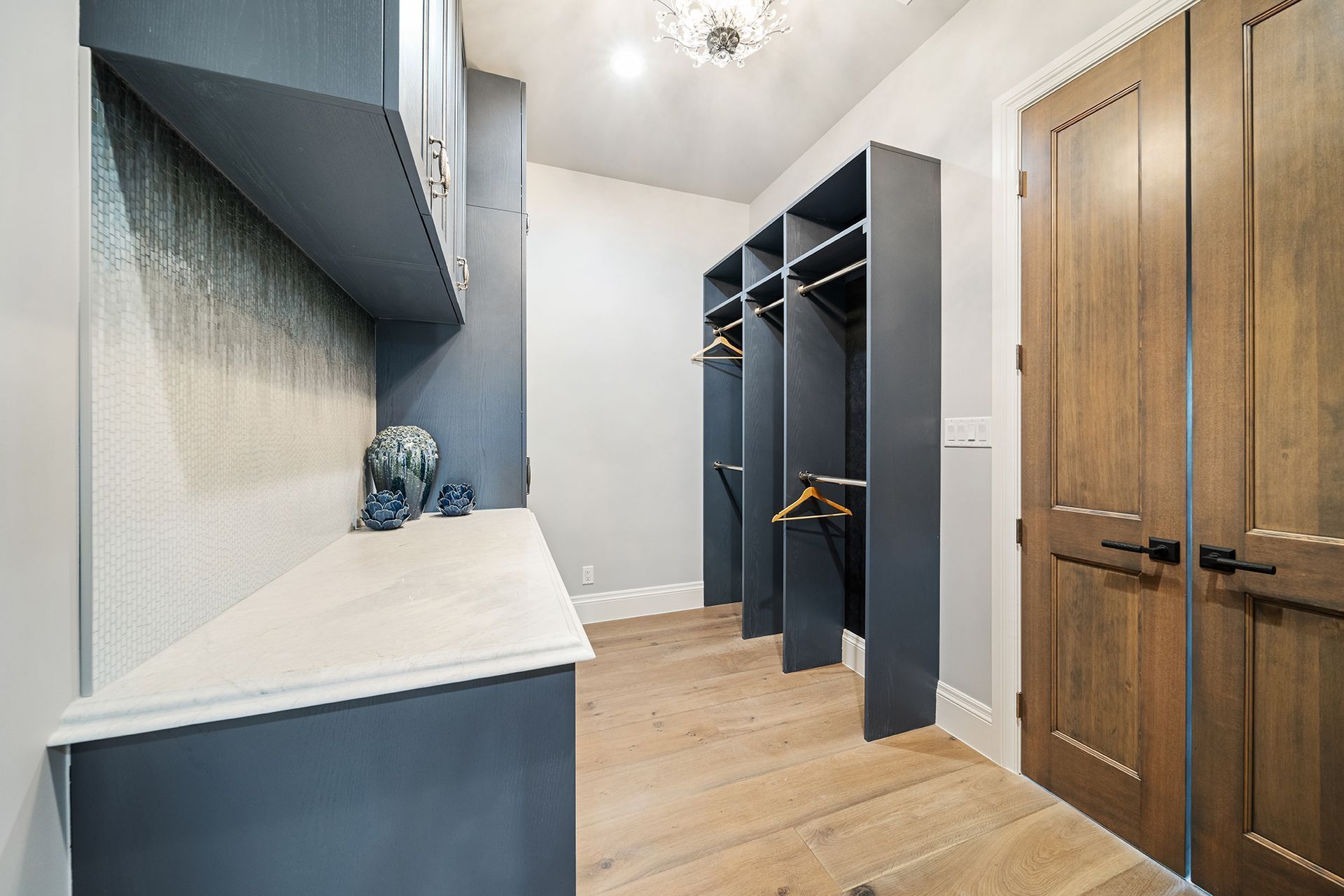 A modern walk-in closet with gray built-ins, wooden doors, and light wood flooring. There are hanging clothes and decorative items.
