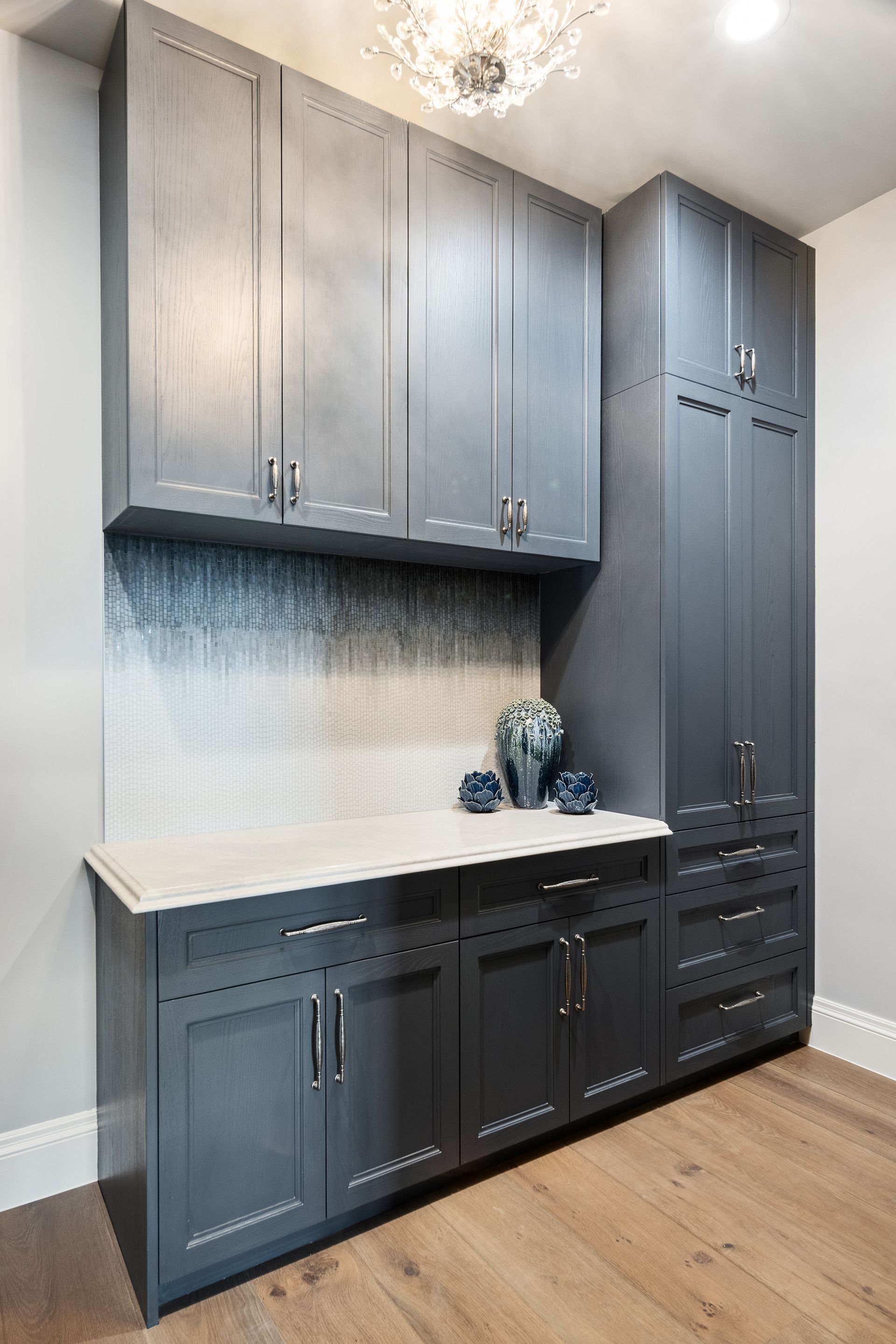 Gray built-in cabinets in a room. White countertop, backsplash, and silver hardware. Wooden floor.