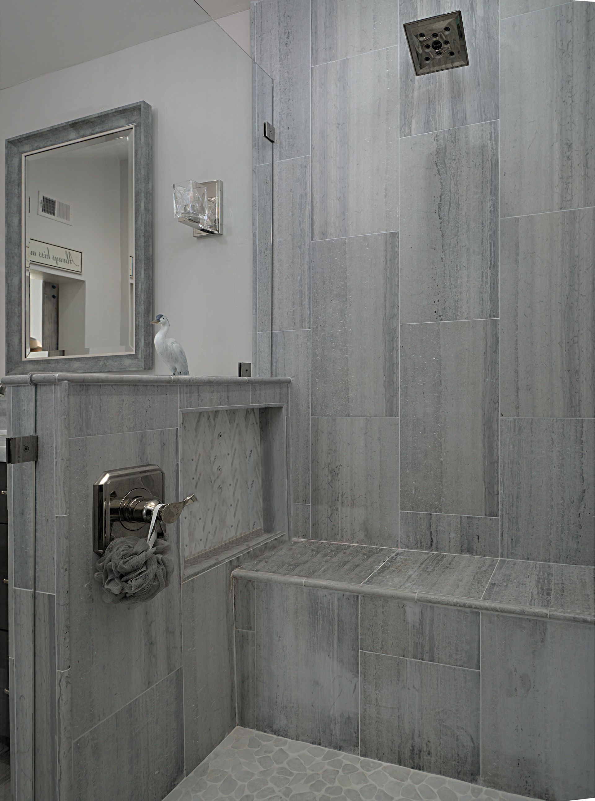 Gray tiled shower with a built-in bench, faucet, and a recessed shelf. A showerhead and glass door are visible.