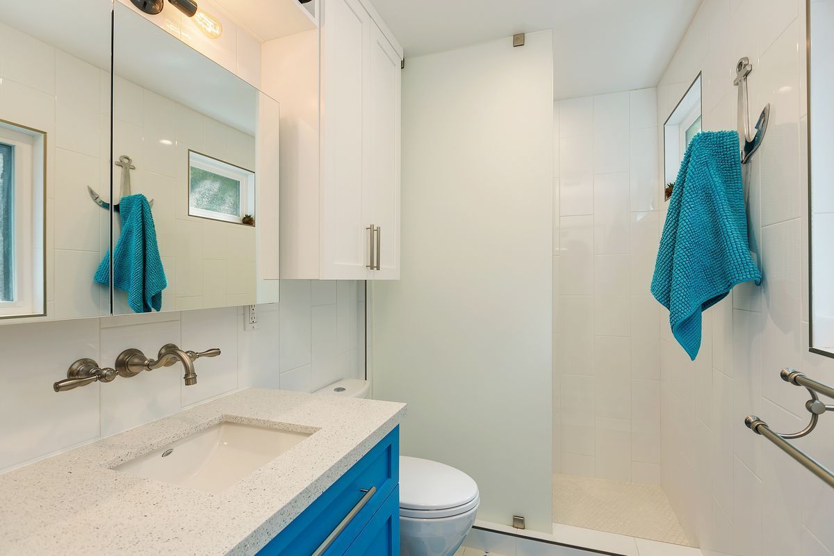 Small, modern bathroom with a blue vanity, white countertop, and glass shower. A turquoise towel hangs on a wall hook.