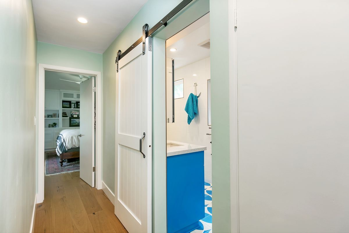 Hallway with a barn door leading to a bathroom and a door to a bedroom. Pale green walls and wood floors.