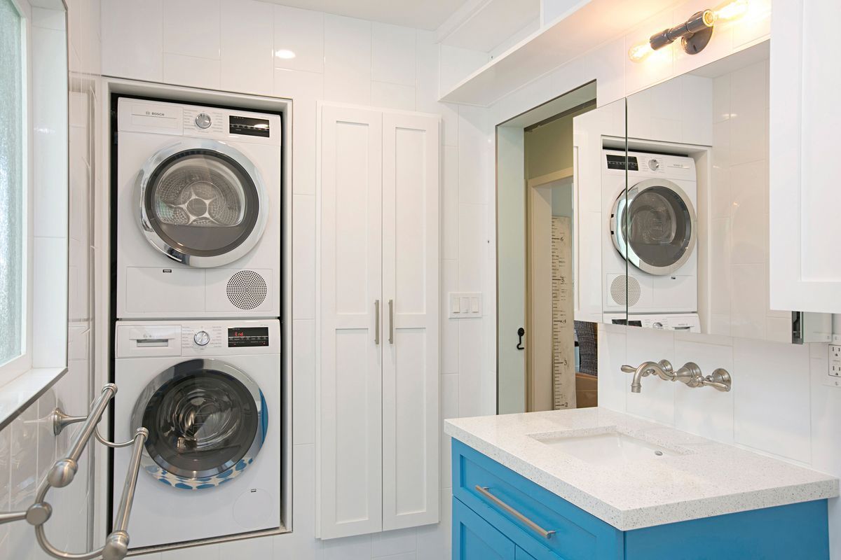 A small, bright bathroom with a stacked washer and dryer, a blue vanity, and white cabinets.