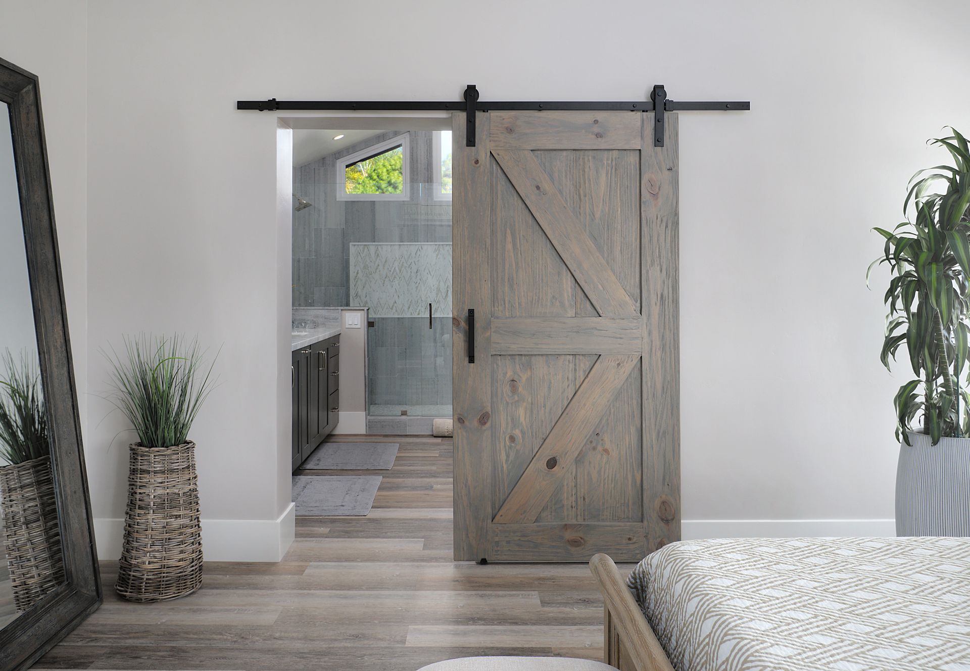 A barn door partially open to a bathroom with a gray-tiled shower, in a bedroom with light wood floors.