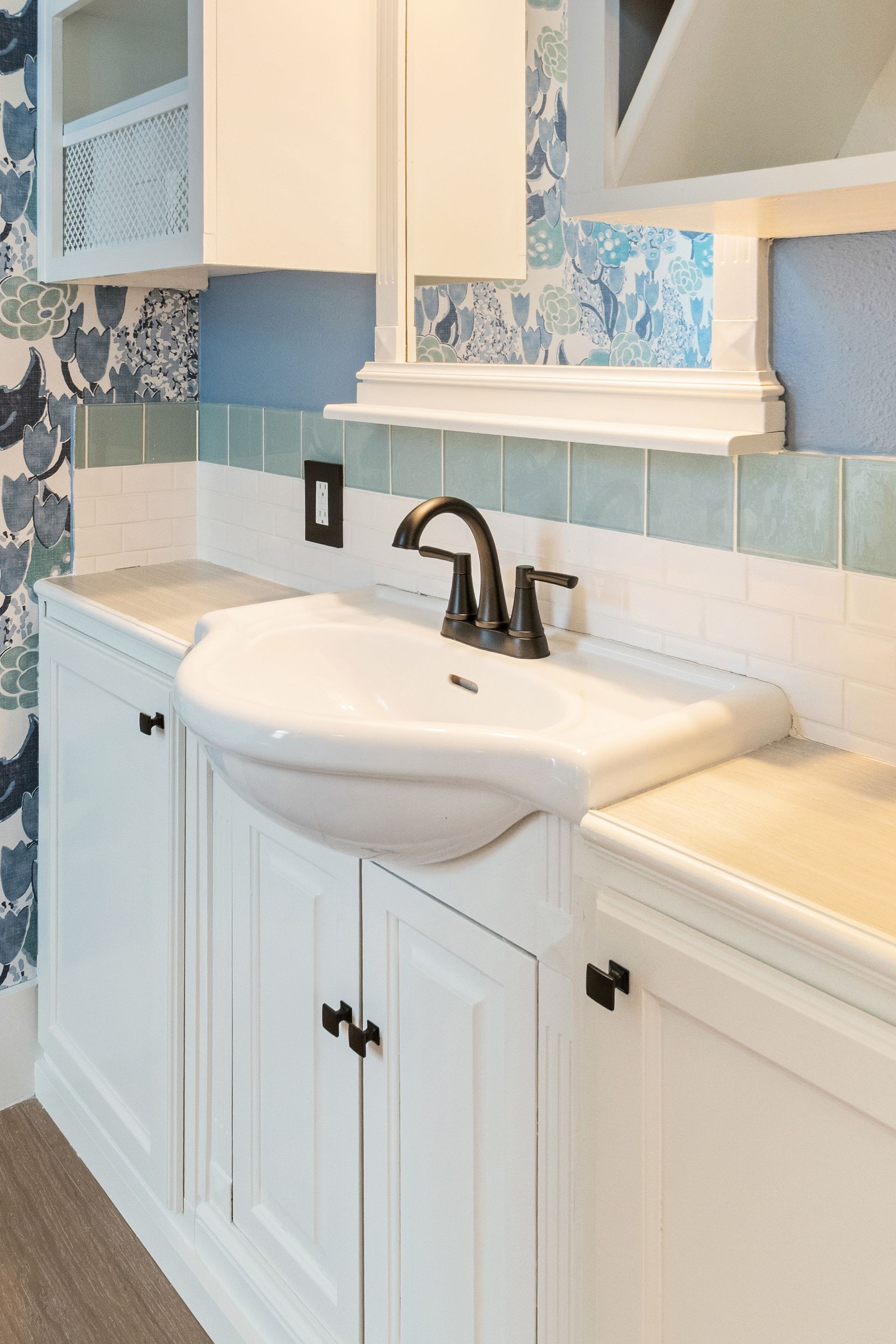 White sink with black faucet in a bathroom with blue patterned wallpaper and white cabinetry.