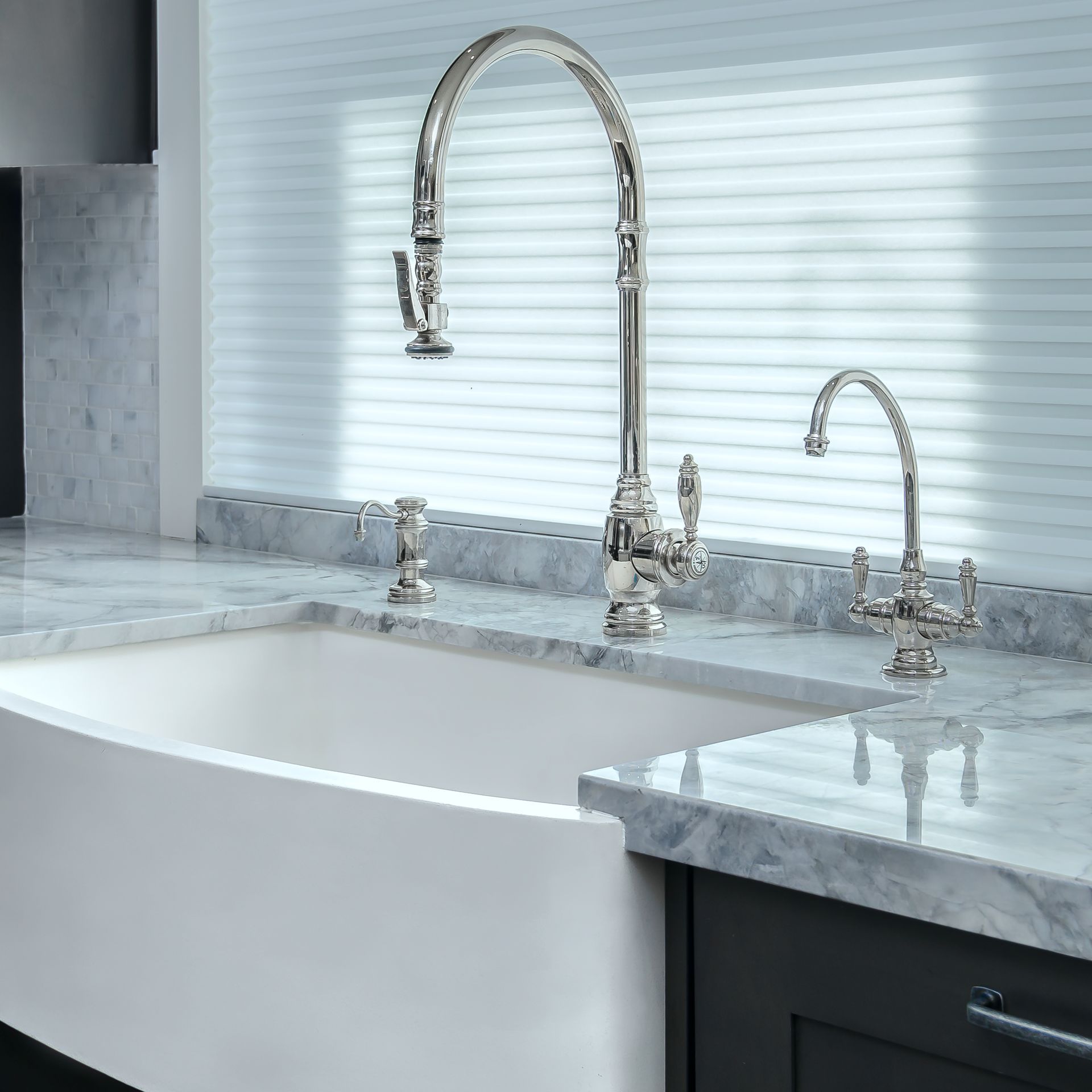 A kitchen sink with a high-arched faucet and a water filter faucet against a marble countertop and a window with blinds.