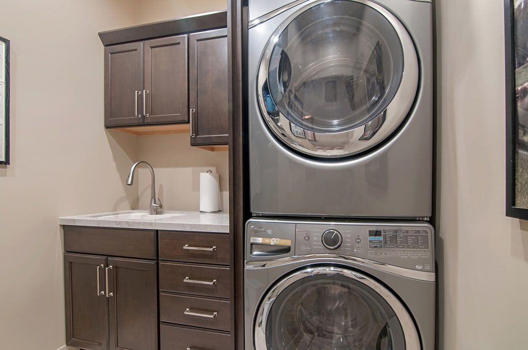 A laundry room with stacked silver washer and dryer, brown cabinets, and a sink.