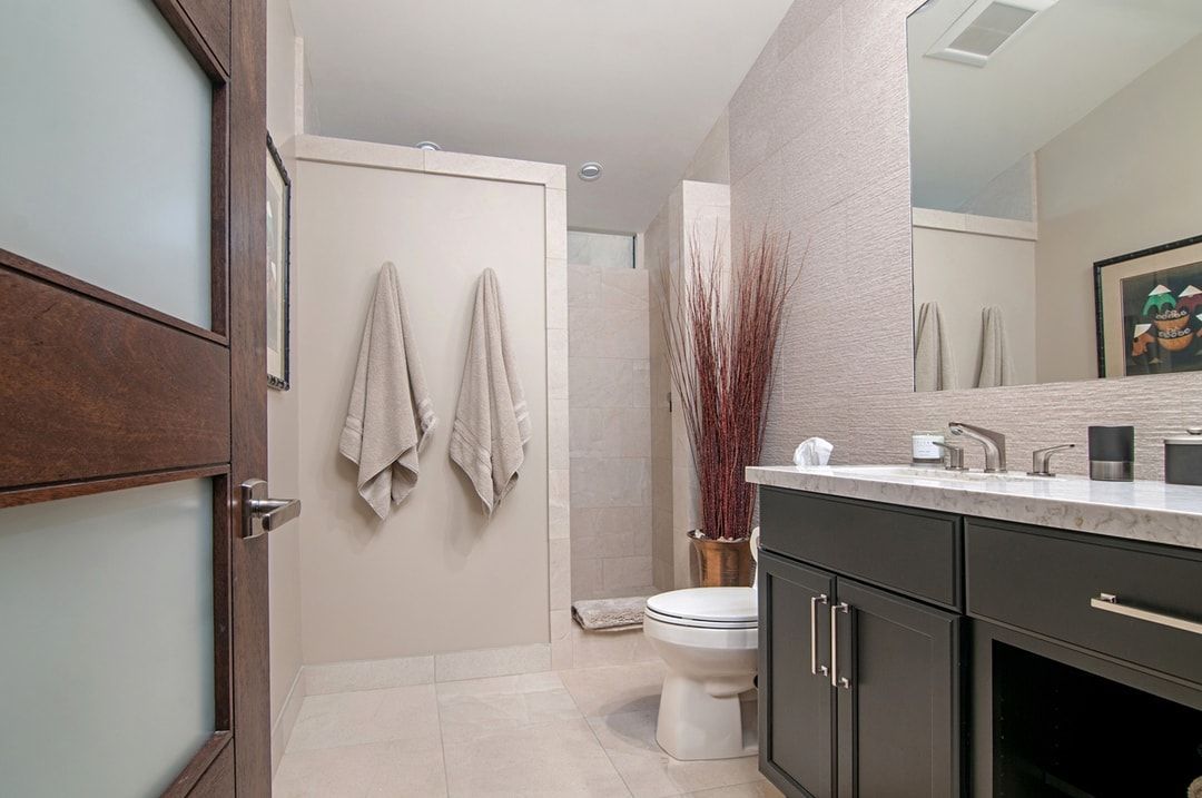 Bathroom with dark cabinetry, a toilet, and a shower. Two tan towels hang on a wall.  The setting has beige and white tiles.