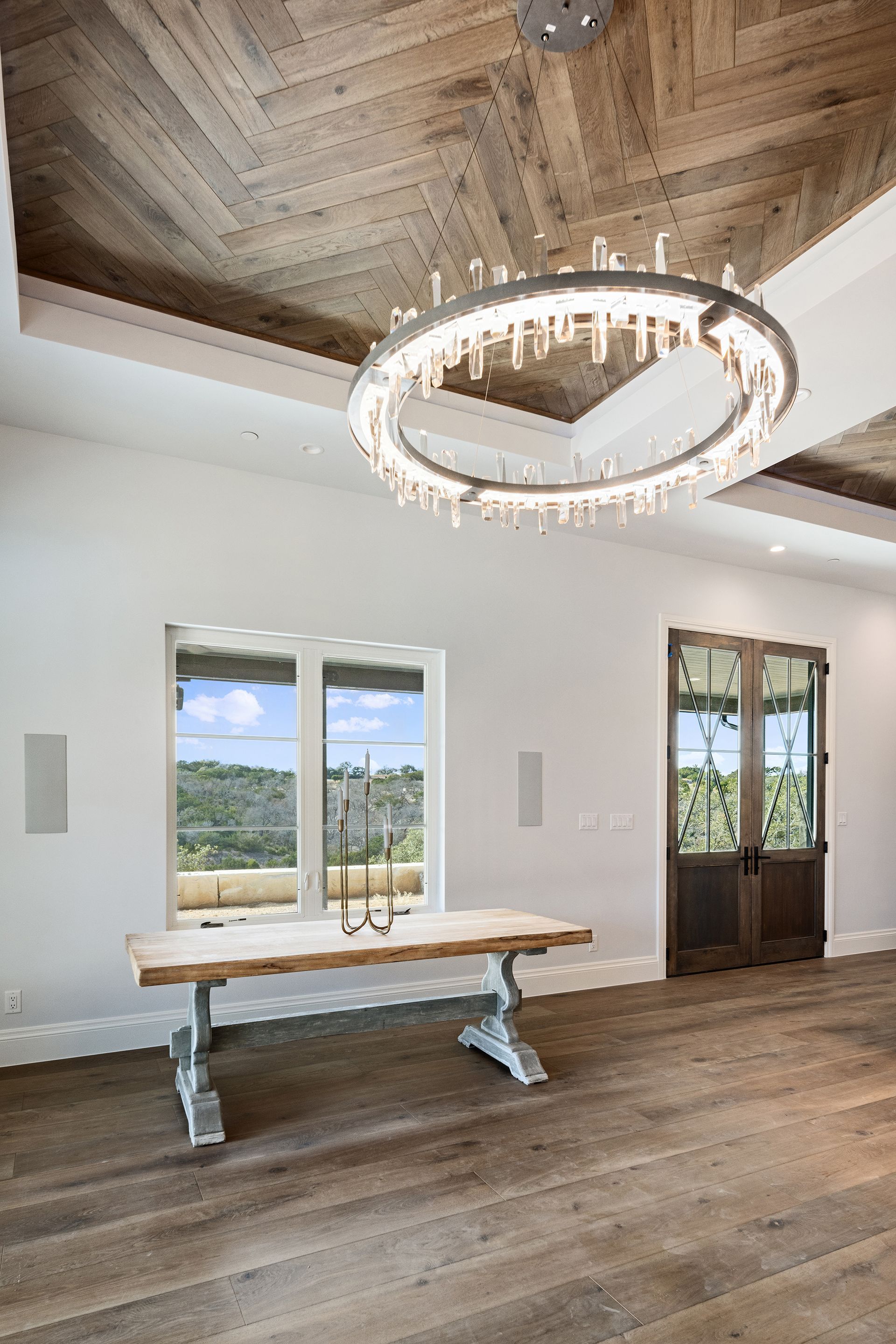 Spacious room with a wooden chevron ceiling, large chandelier, windows, and a rustic table. A door and a light-colored hardwood floor are also present.