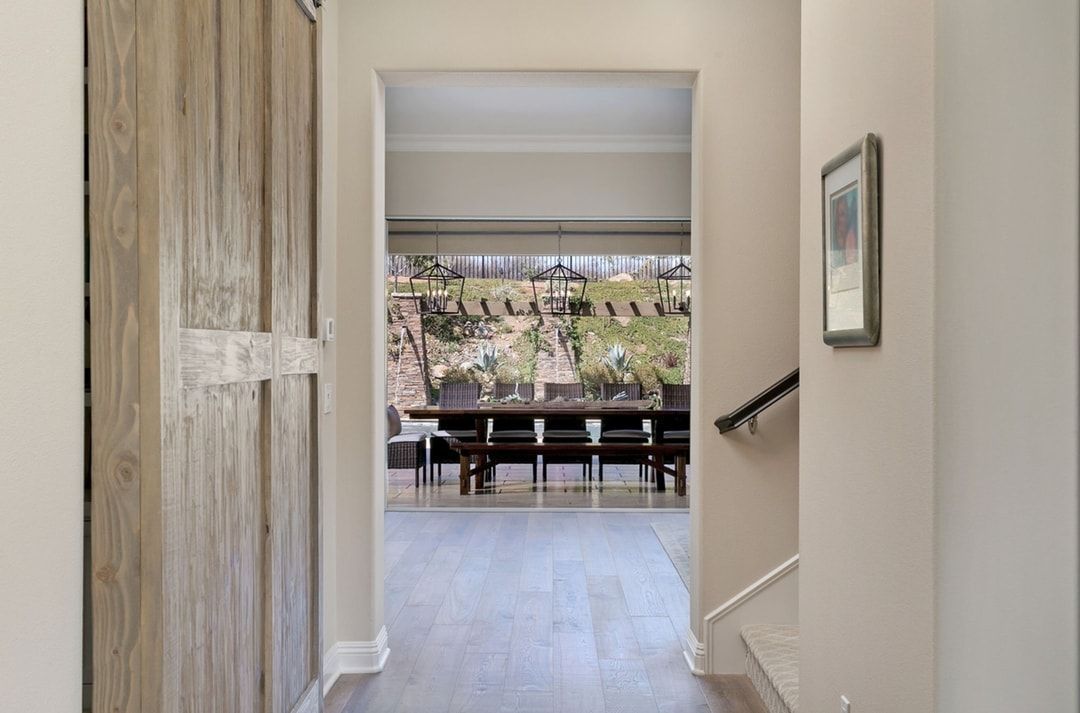 Hallway with wood door on left, leading to dining room with large table and view of landscape. Stairs and artwork on right.