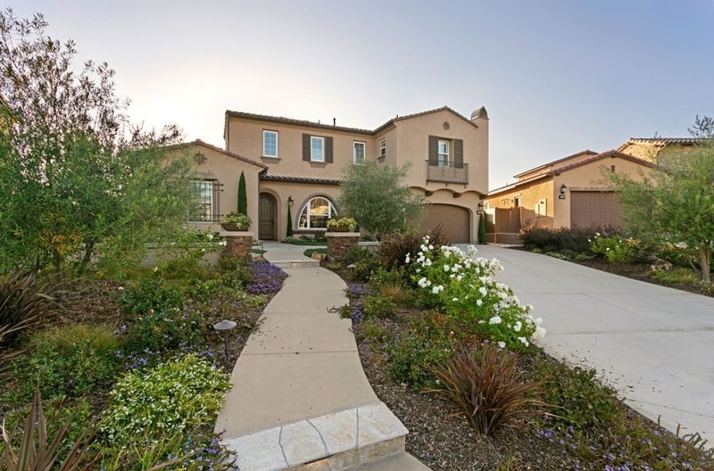 Two-story beige house with a pathway leading to the front door; surrounded by lush landscaping and a driveway.