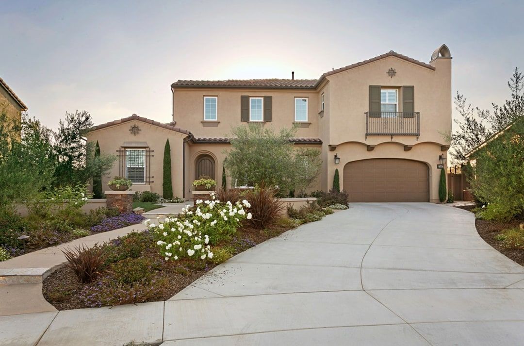 Beige two-story house with a curved driveway, featuring a garage and landscaping.