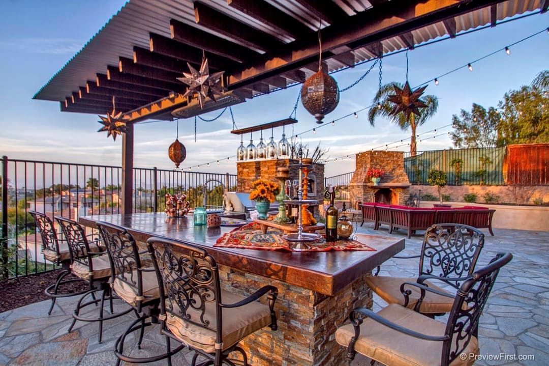 Outdoor bar with stone facade and wooden countertop, surrounded by ornate metal bar stools, under a wooden pergola strung with lights, overlooking a sunset view.