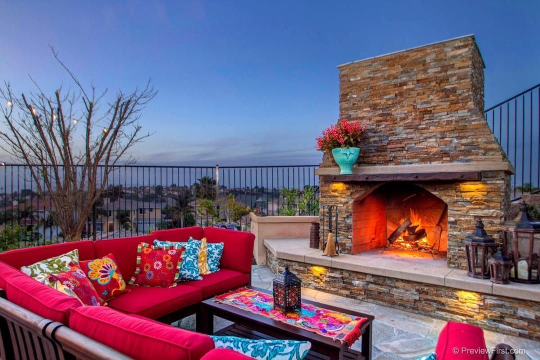 Cozy outdoor patio with a stone fireplace, red sofa, colorful cushions, and a view at dusk.