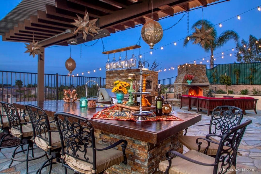 Outdoor patio with a bar, fireplace, and seating, illuminated by string lights and hanging lanterns against a twilight sky.