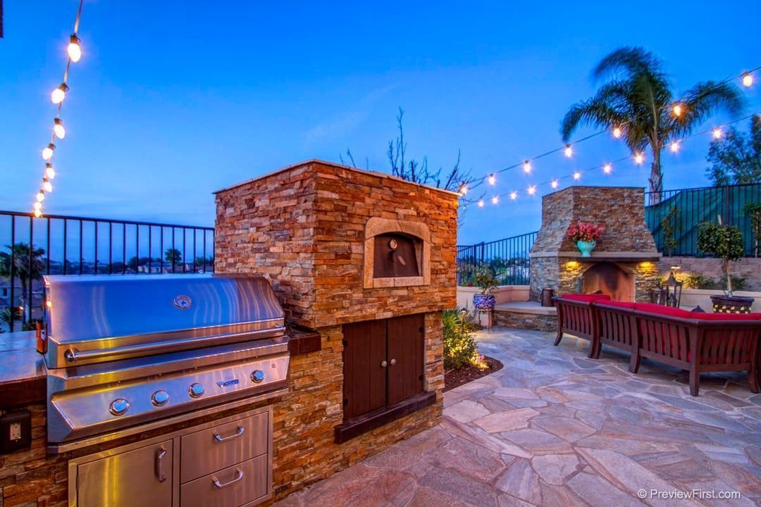 Outdoor kitchen with a grill, brick oven, and fireplace, illuminated by string lights, set against a twilight sky.