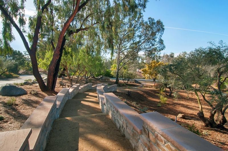 Stone pathway winds through a sunny park with trees and low walls. The sky is bright blue.