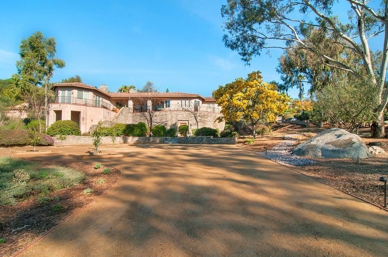 A large, two-story house with a light-colored facade sits behind a long dirt driveway, surrounded by landscaping under a bright blue sky.