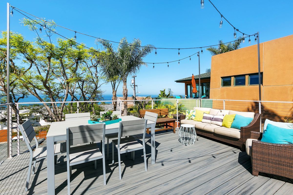 An outdoor deck with a dining table and seating area, strung with lights, overlooking a coastal view.