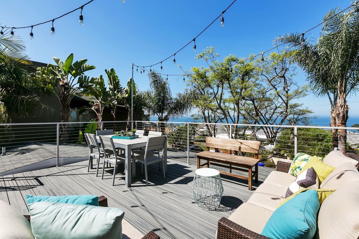 A sunny outdoor deck with a dining table, seating area, and ocean view. String lights are strung overhead, with trees and blue sky in the background.