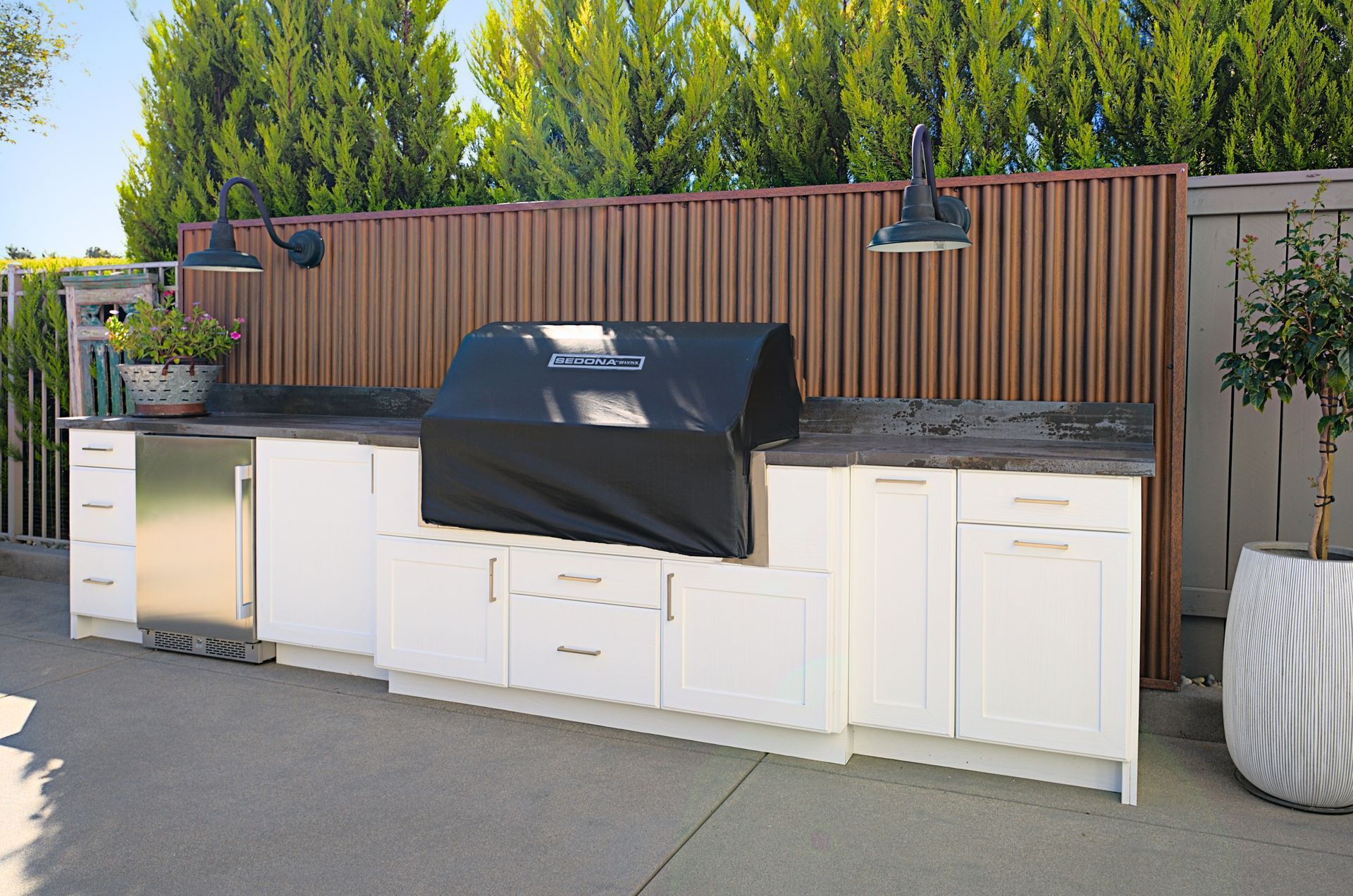 An outdoor kitchen with white cabinets, a covered grill, and a brown wooden privacy screen against a backdrop of green trees.