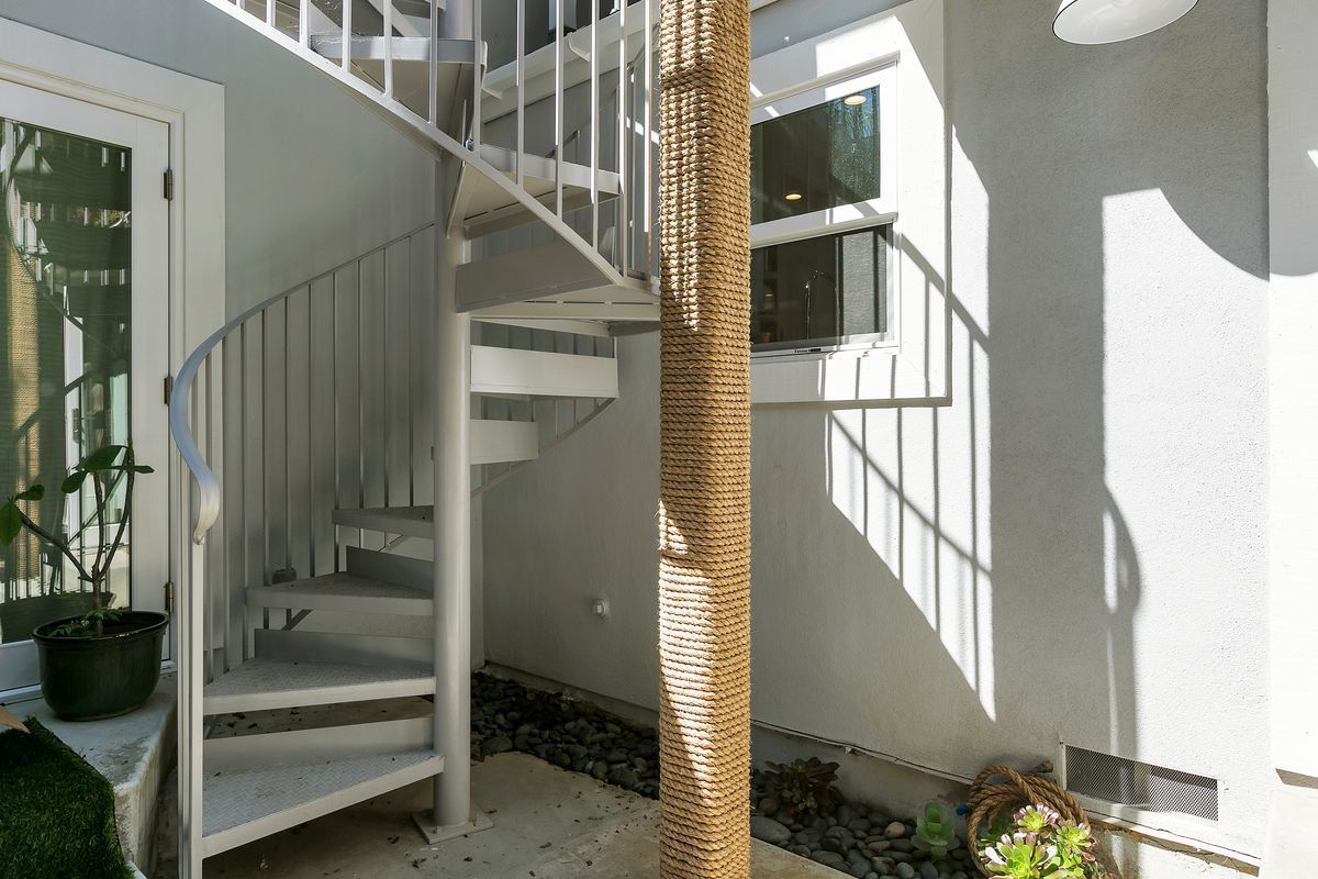 White spiral staircase outdoors next to a textured, brown post and a white wall, with a door and a window visible.