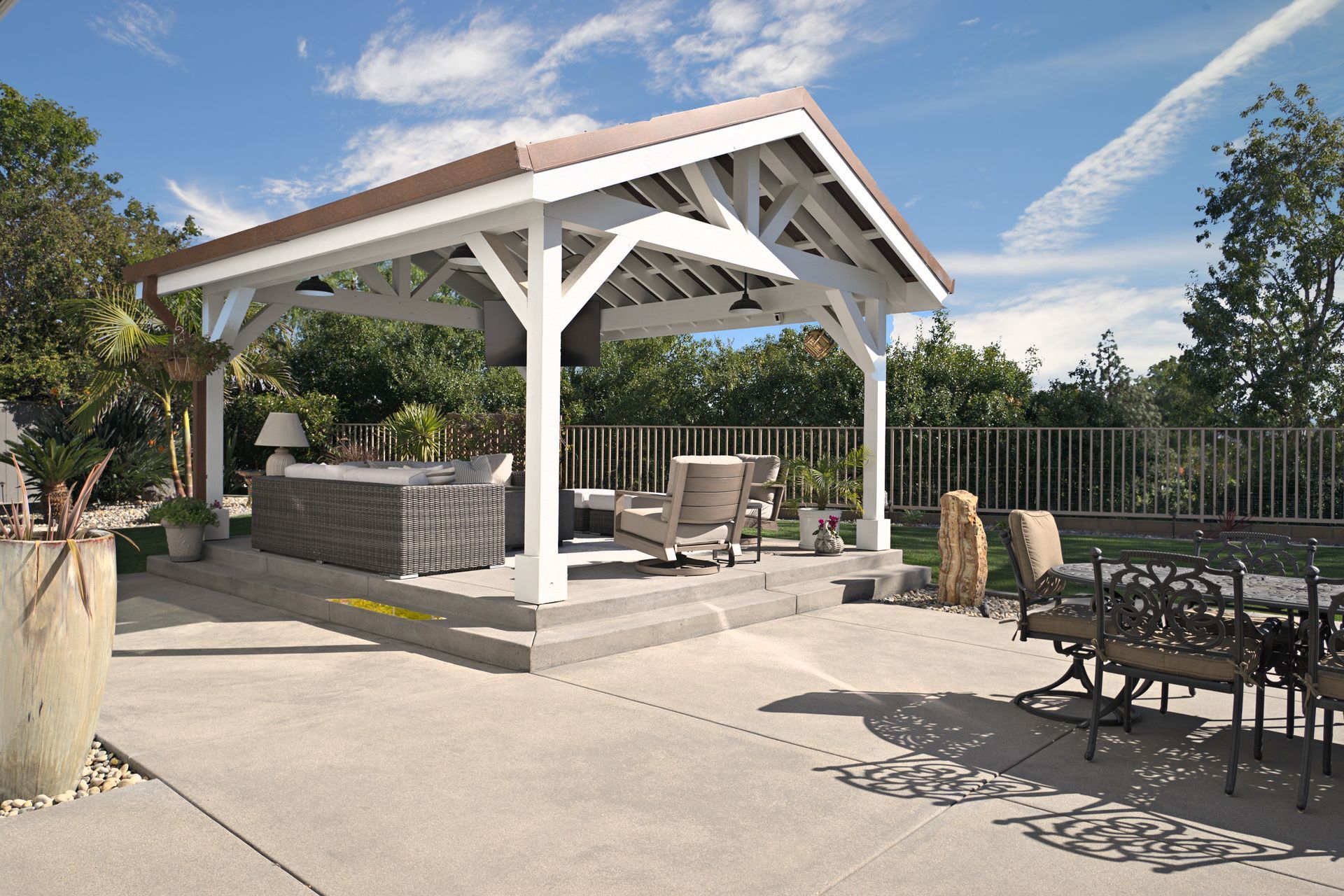 Outdoor patio with a white gazebo over a seating area and dining table set. Blue sky with clouds in the background.