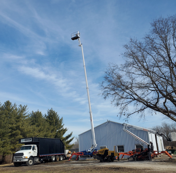 Truck with lift arm extended towards a building for work. White truck, blue sky, bare tree.