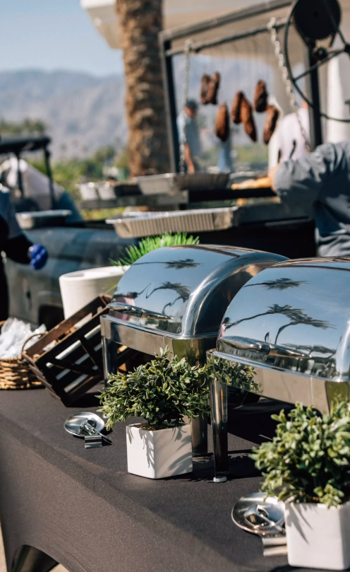 A buffet table with stainless steel trays and potted plants on it.