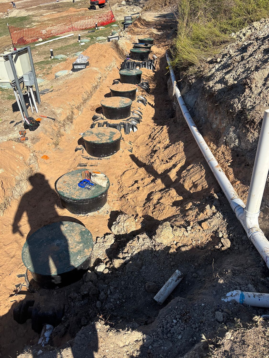 A row of round, dark-colored septic tank access covers installed in a trench on a dirt construction site.