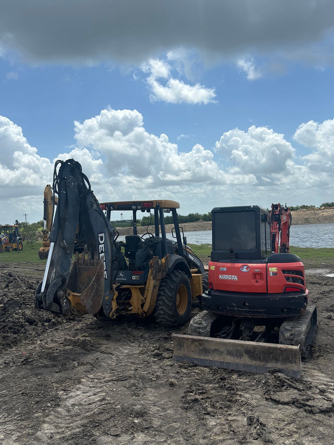 A yellow John Deere backhoe and a red excavator parked in a muddy field under a cloudy blue sky.
