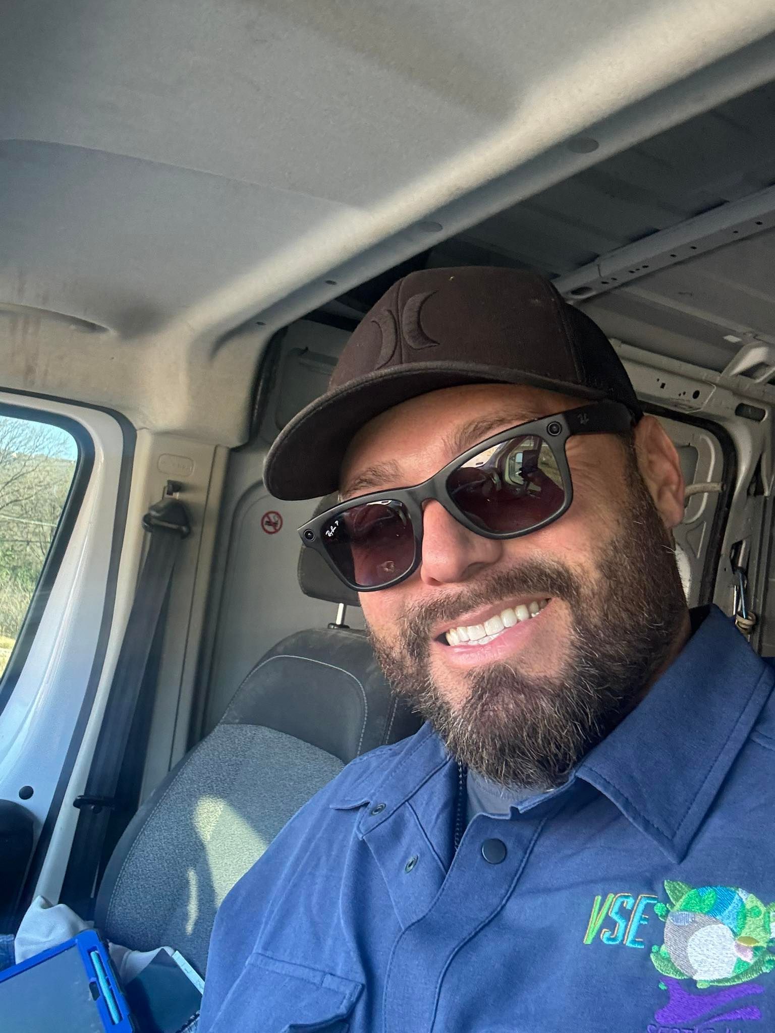 A smiling person with a beard, wearing sunglasses and a baseball cap, sits in the driver's seat of a work van.