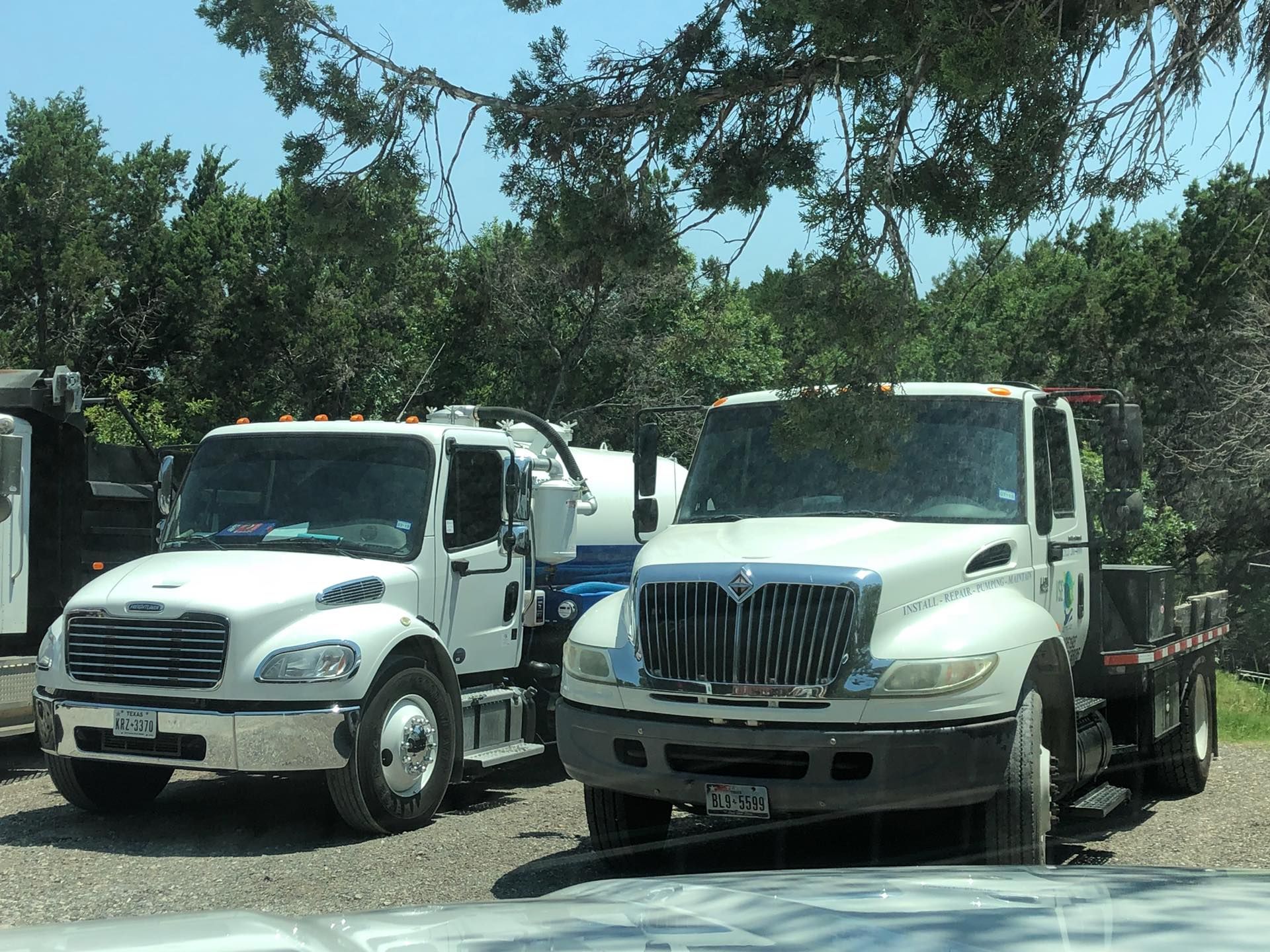 Two white commercial trucks parked side-by-side on a gravel lot under trees.