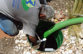 A technician services an outdoor septic or pump system using a green suction hose.