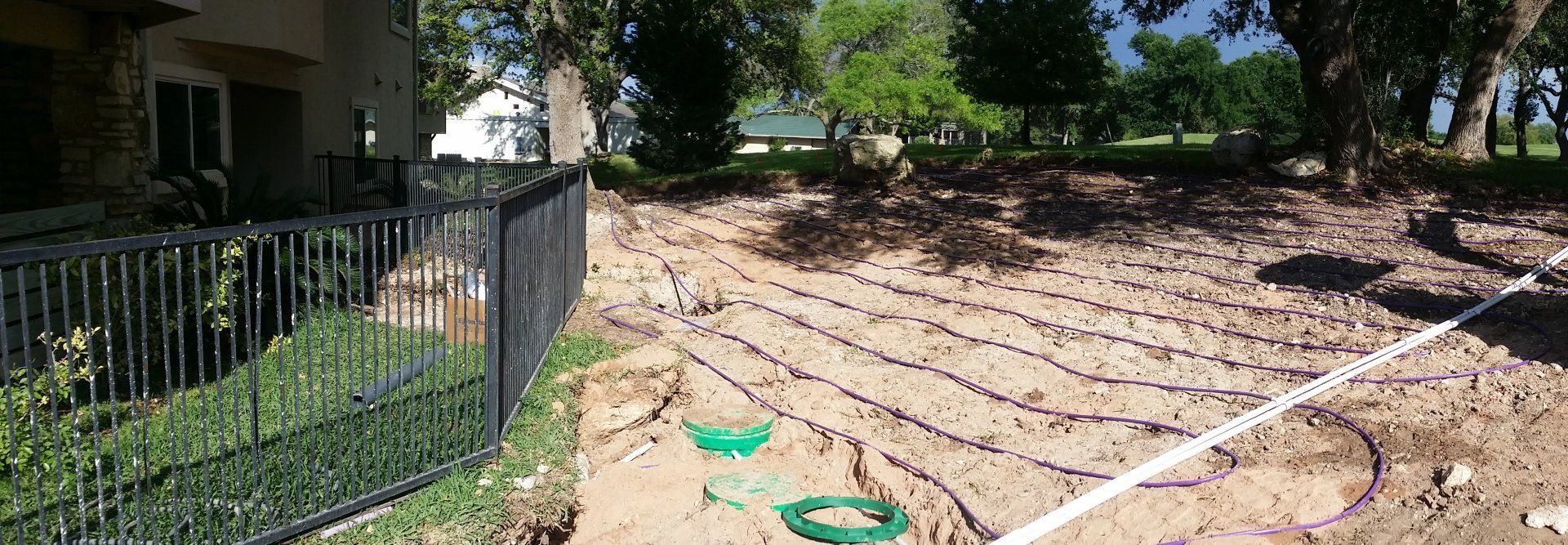 Drip irrigation lines laid out across a residential yard next to a metal fence and septic tank access covers.