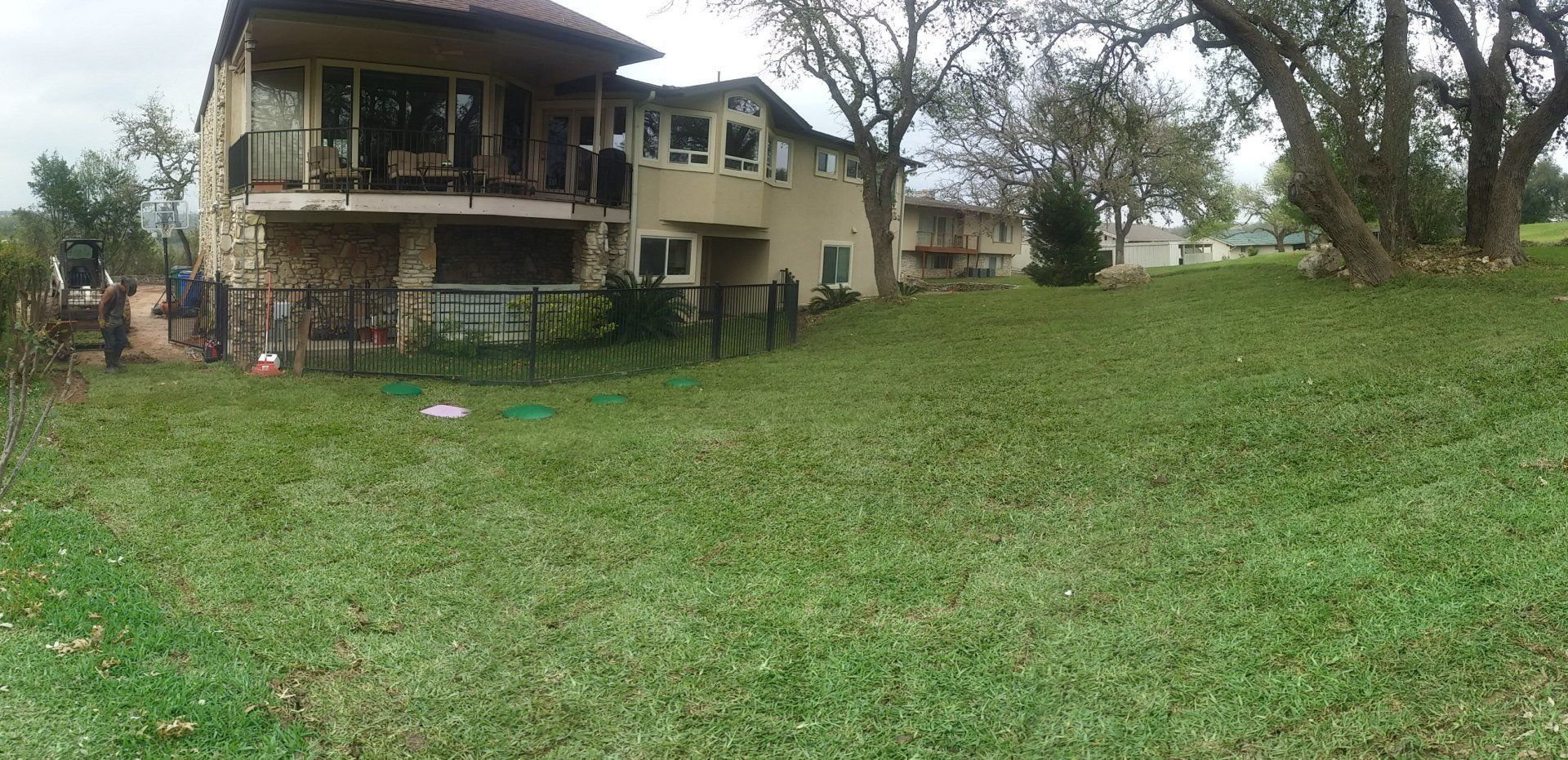 A two-story tan house with a balcony and a fenced yard sits on a grassy hill next to a large tree under an overcast sky.