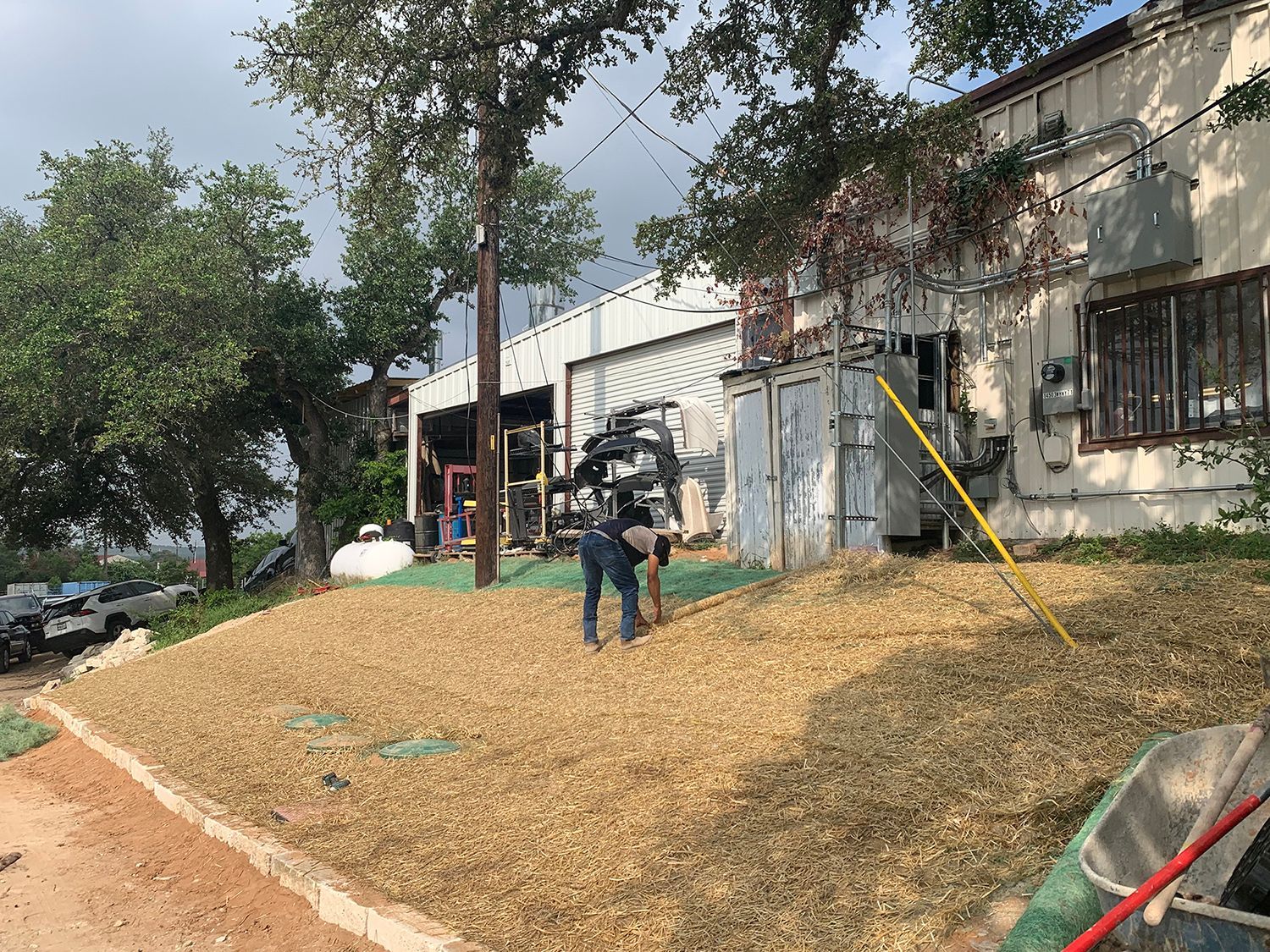 A person works on a sloped, wood-mulched area next to a building and trees on a sunny day.