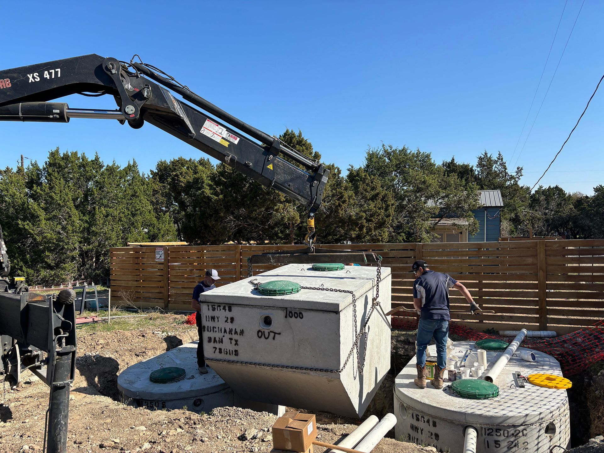 An excavator lowers a large concrete septic tank into a hole next to a worker near a wooden fence on a sunny day.