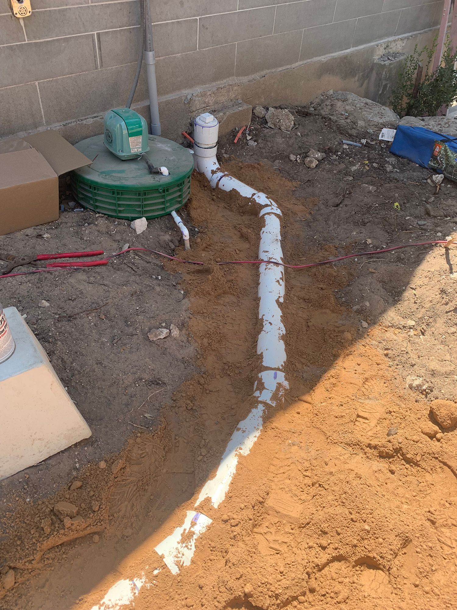 White PVC piping laid in a shallow dirt trench in a backyard, connecting to a green drainage catch basin.
