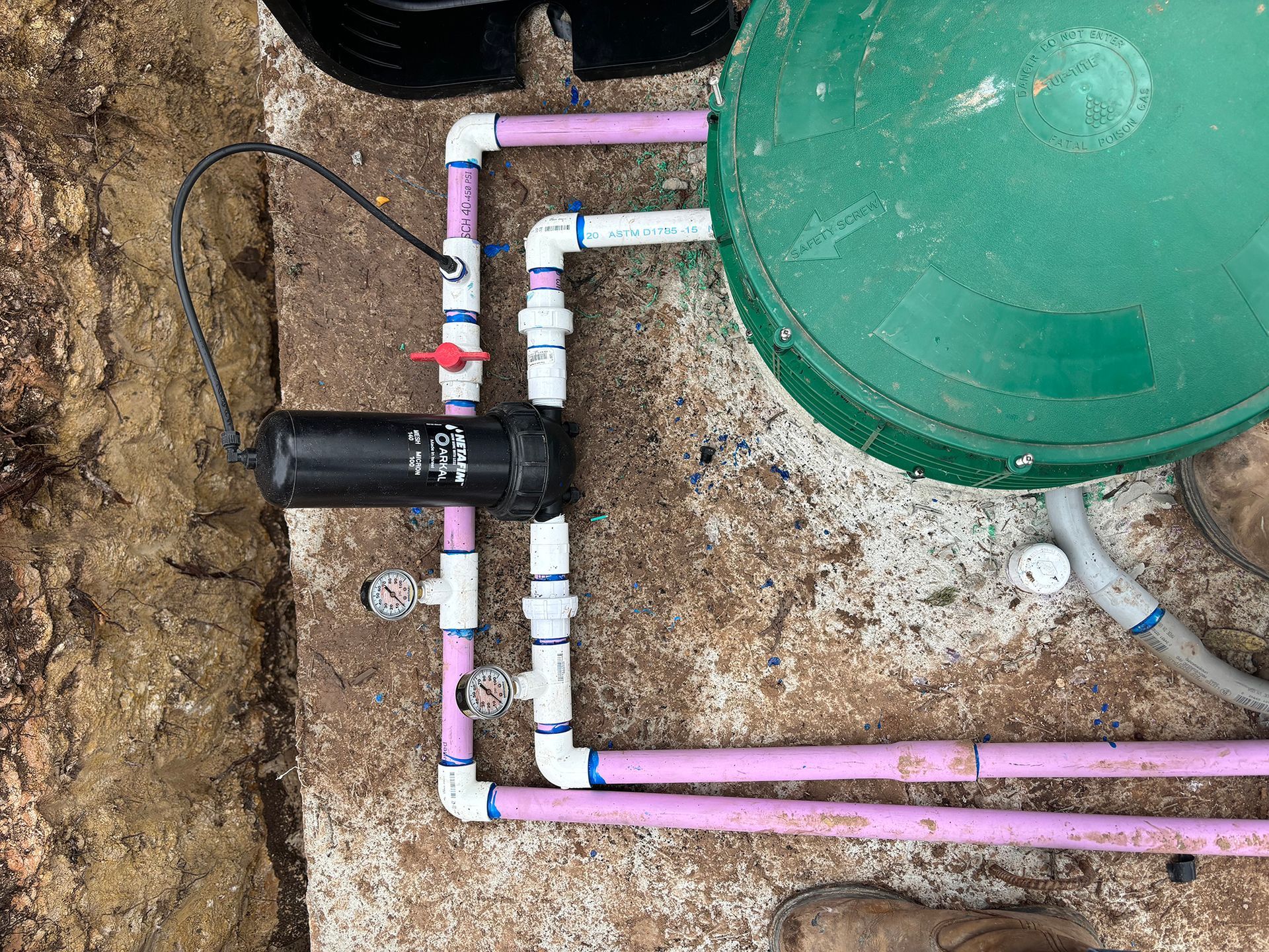 Irrigation plumbing featuring a black filter, pressure gauges, and purple PVC pipes connected to a green tank.
