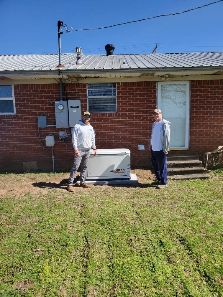Two people standing beside an outdoor generator in front of a brick house on a sunny day.