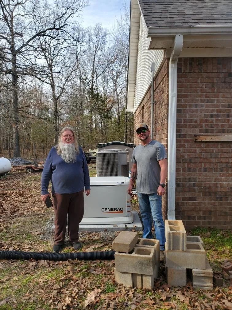 Two men stand beside a house and stacked cinder blocks near an outdoor HVAC unit.