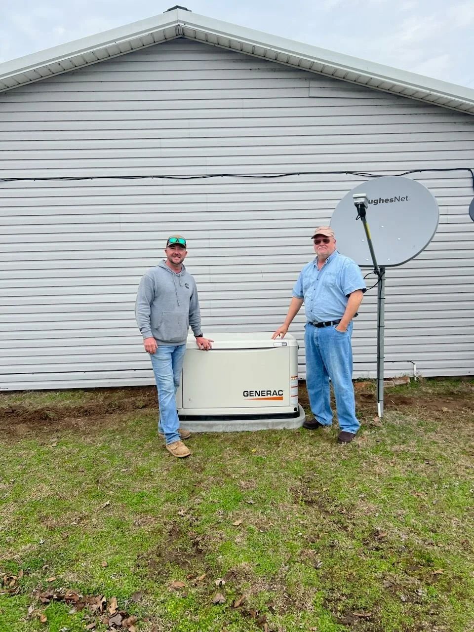 Two people stand beside an outdoor generator against a white house, with a satellite dish nearby.