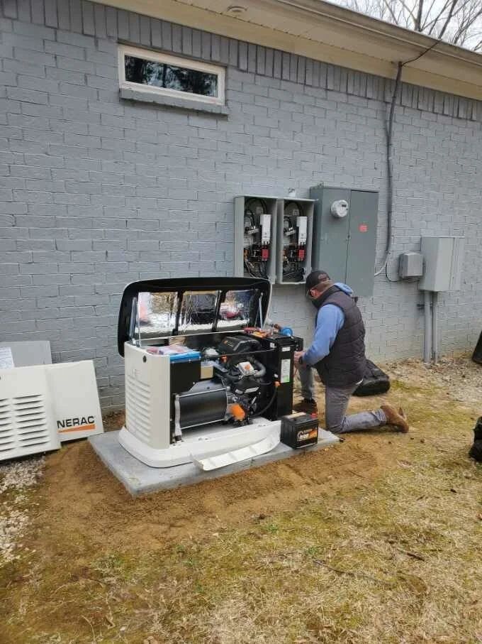 Technician kneeling beside an outdoor generator beside a gray building, inspecting its open panel.