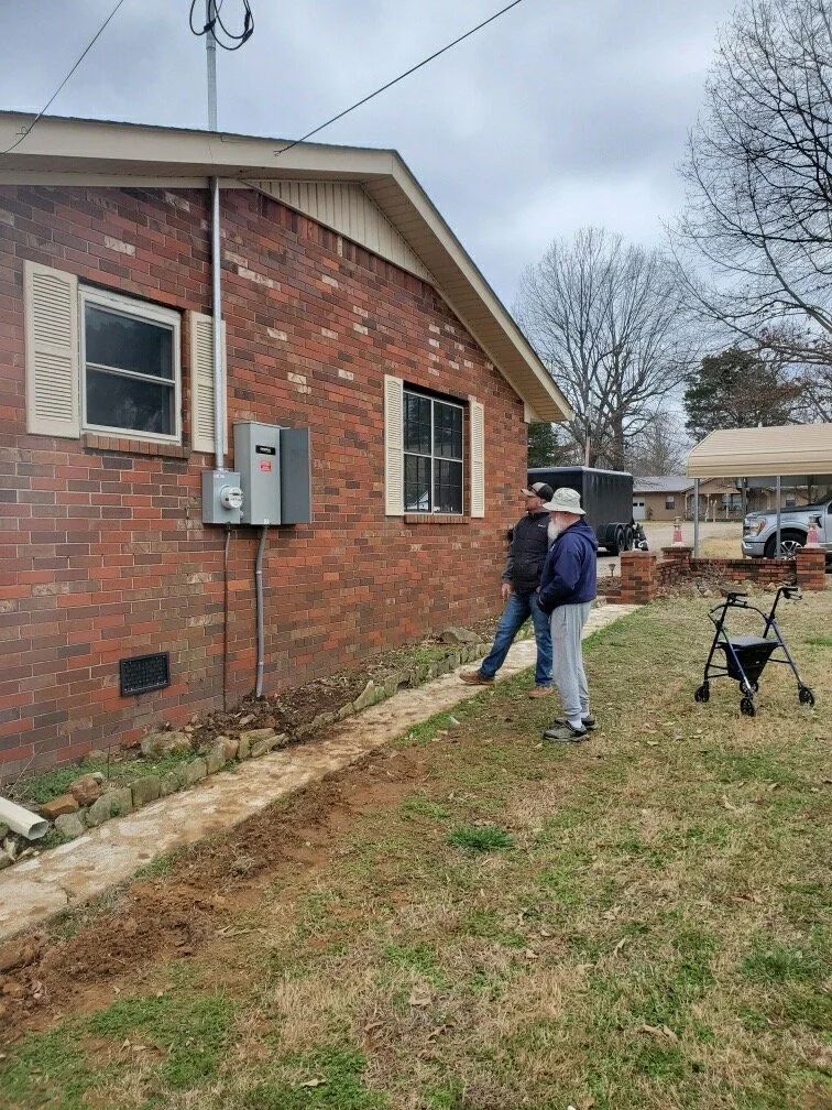 Two people working outside a brick house, one using a blower along the foundation in a grassy yard.