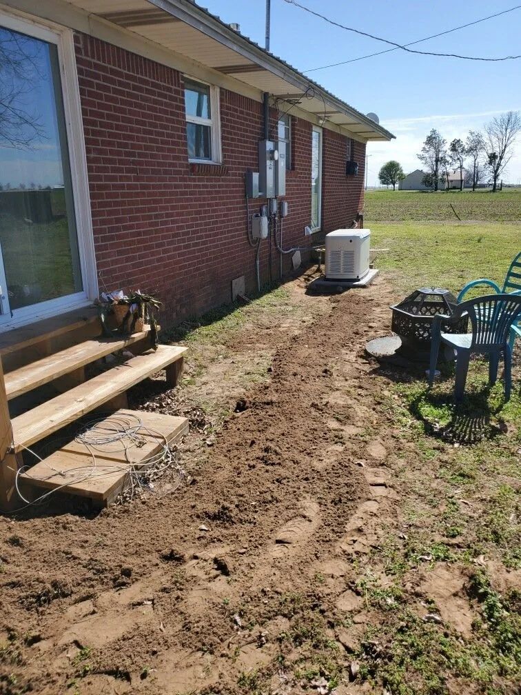 Side yard beside a brick house, with a dirt trench, patio chairs, and an HVAC unit in the distance.