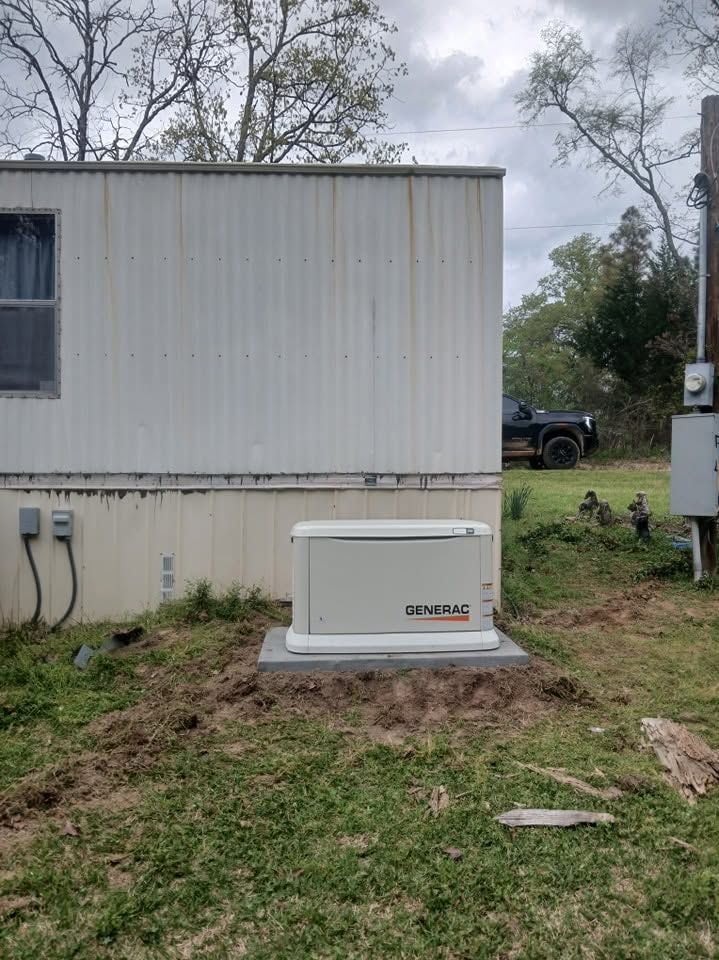 White mobile home with a beige utility unit in a grassy yard, trees and a parked truck in the background