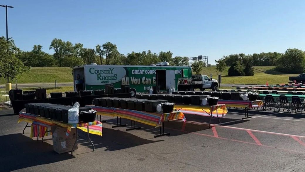 Outdoor event booths under tents with tables and signs in a sunny parking lot, with trees and a trailer in back